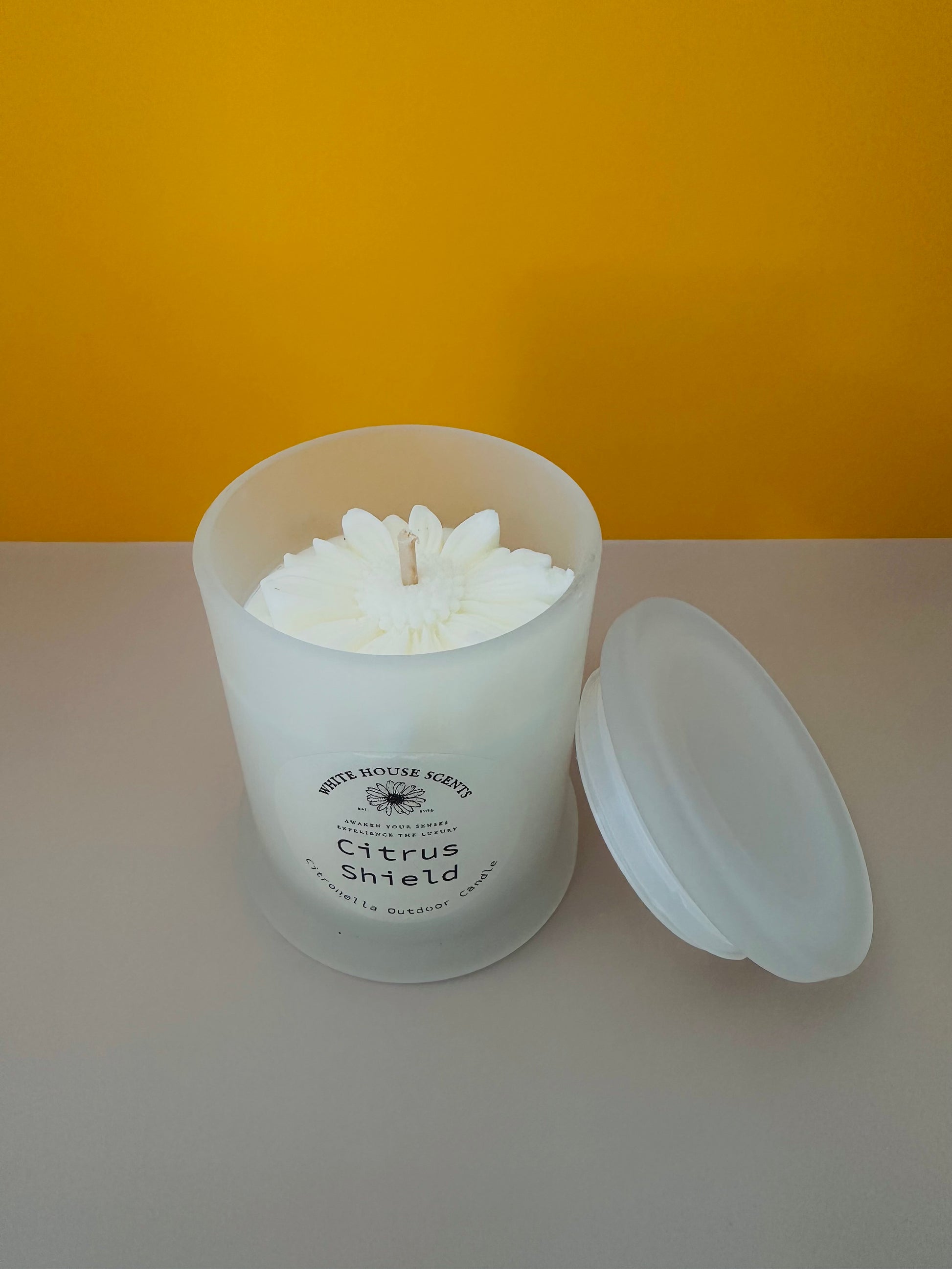 White candle in a glass jar with a label on a white surface and yellow background